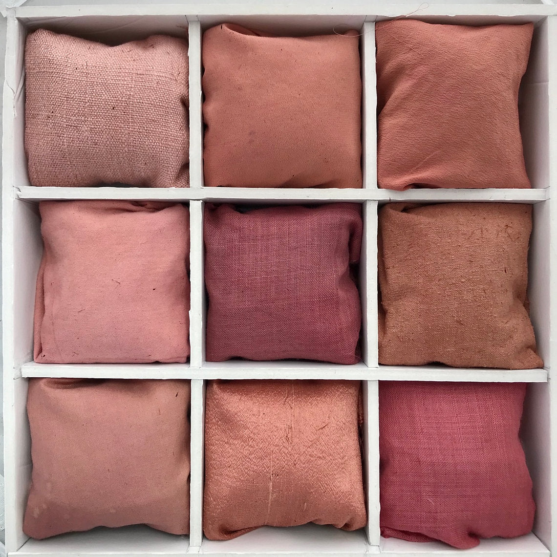 Nine pink fabrics dyed with Alkanet Roots (Alkanna tinctoria) arranged in a 3x3 grid on a white wooden shelf.