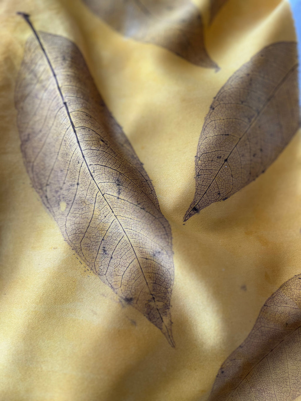 A close-up of a yellow fabric printed with realistic brown dried leaves, showing the fine leaf vein patterns.