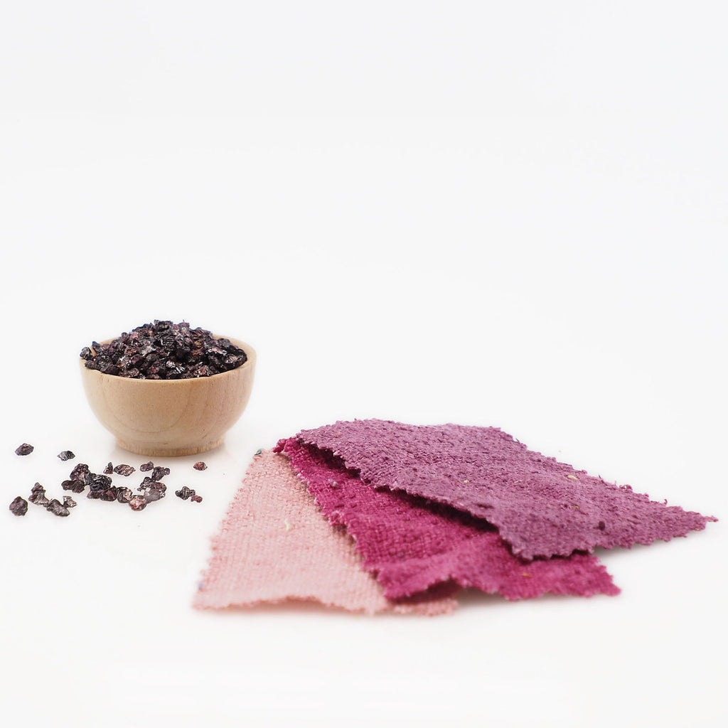 Three purple fabric samples on a white background with a small wooden bowl of dark berries.