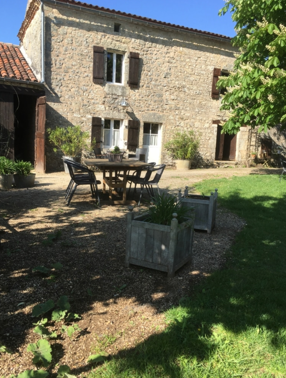 A stone farmhouse courtyard with a wooden dining table and chairs, potted plants, a gravel and grass yard, and a tree in the foreground.