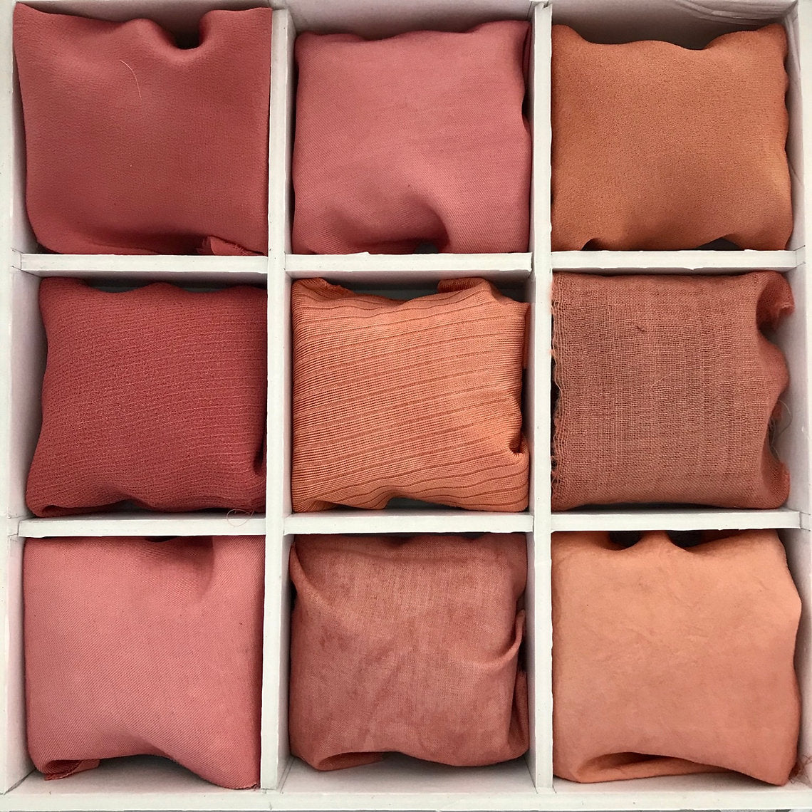 Nine square pillows in various shades of red and orange dyed with Madder Extract (Rubia Tinctorum) arranged in a grid on a white background.