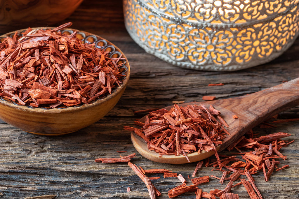 Dried red herbs in a wooden bowl and on a wooden spoon with a decorative metal container in the background.