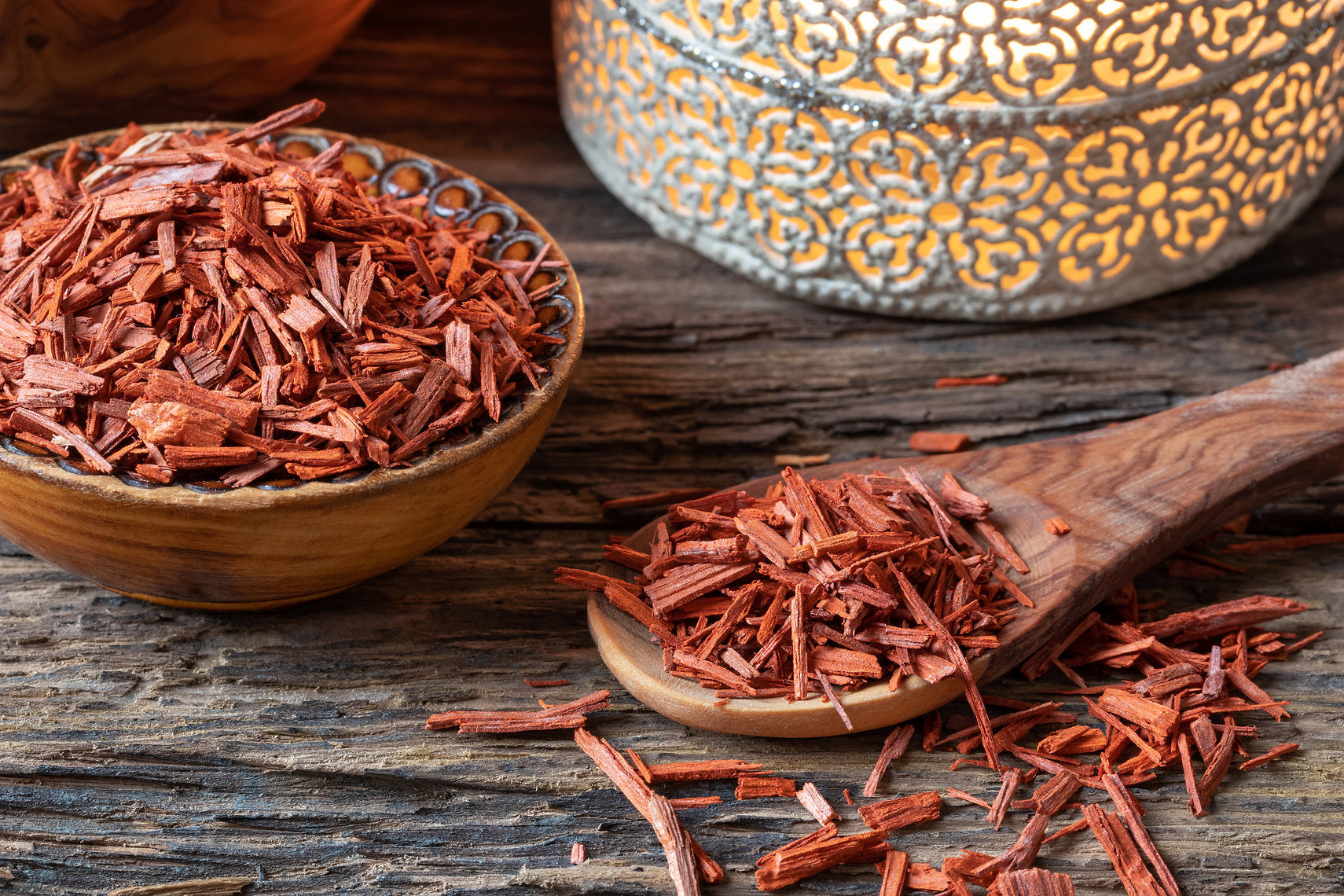 Dried red herbs in a wooden bowl and on a wooden spoon with a decorative metal container in the background.