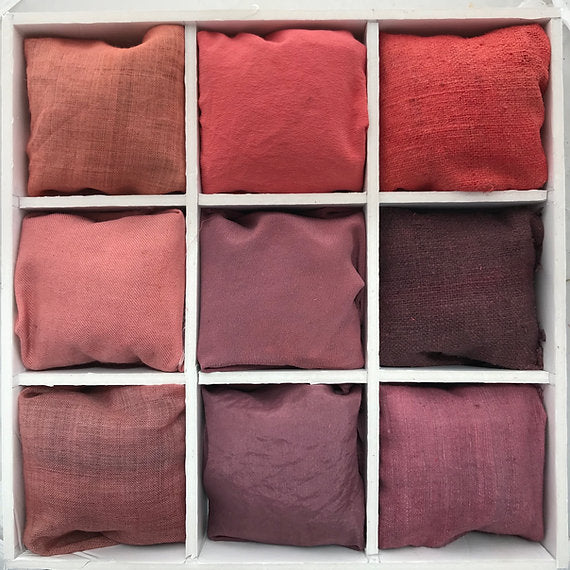 Nine fabric pillows in various red and pink shades arranged in a grid on a white wooden shelf.