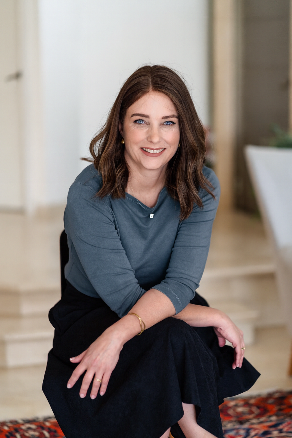Suzanne Dekel sitting on a rug indoors wearing a blue top and black skirt.